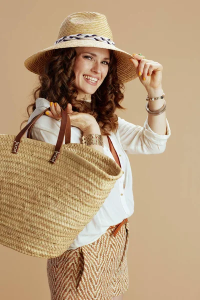 Plaj tatili Beyaz bluz ve şortla bej arka planda hasır çanta ve hasır şapkayla gülümseyen 40 yaşındaki zarif ev hanımı..