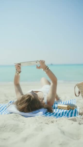 ストローバッグで海岸のリラックスしたエレガントな女性とタブレットPcの空白の画面を示すストライプタオル — ストック動画