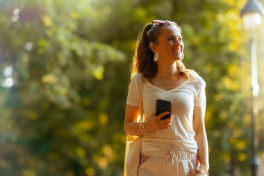 Yaz zamanı. Beyaz tişörtlü, çantalı mutlu orta yaşlı kadın akıllı telefon kullanıyor ve parkta yürüyor..