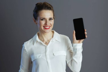 Beyaz bluzlu 40 yaşındaki mutlu stil sahibi kadın çalışanı gri arka planda akıllı telefon boş ekranı gösteriyor..