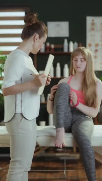 医療時間について クリップボードと十代のクライアントが足の痛みの苦情を聞いているマッサージキャビネットの心配マッサージセラピスト女性 — ストック動画