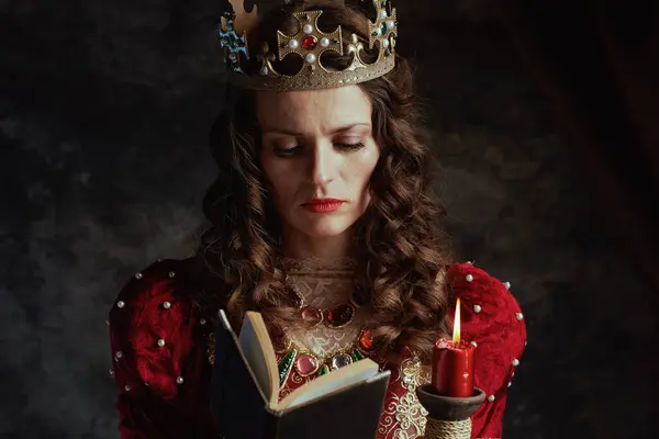 medieval queen in red dress with book, candle and crown on dark gray background.