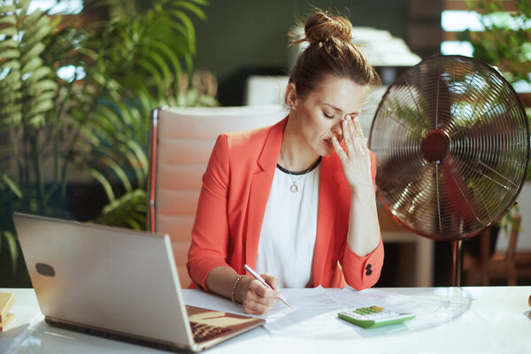 Sustainable workplace. unhappy modern small business owner woman in modern green office in red jacket with electric fan and laptop.