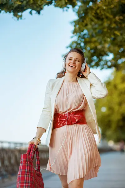 ヘッドフォンとウォーキングで音楽を聞く街のピンクのドレスとホワイトジャケットで幸せなトレンディな女性 — ストック写真