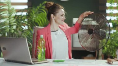Sürdürülebilir iş yeri. Gülümseyen modern iş kadını iş yerinde kırmızı ceketli, elinde bir şişe su ve elektrikli vantilatörle..