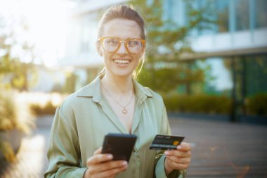 Gülümseyen, orta yaşlı, yeşil bluzlu, gözlüklü, akıllı telefon ve kredi kartlı, iş bölgesinde çalışan bir kadın..