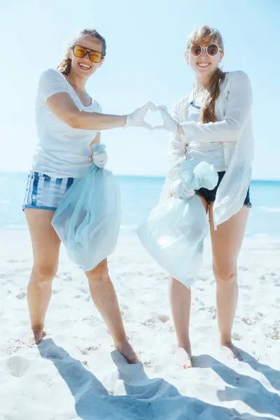 smiling female volunteers on the beach with trash bags collecting trash and showing heart shaped hands.