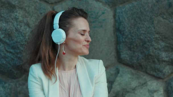 幸せな若い女性 ピンクのドレスと街の白いジャケット 石の壁にヘッドフォン付き — ストック動画