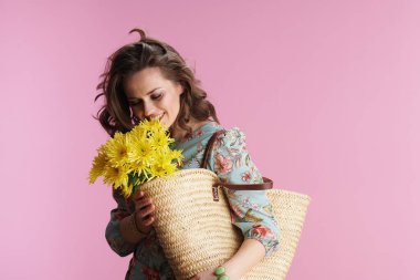 Gülümseyen orta yaşlı kadın, uzun dalgalı esmer saçlı, sarı kasımpatı çiçekli ve pembe arka planda saman çantalı..