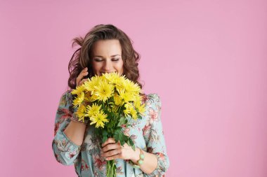 Uzun dalgalı esmer saçlı, sarı kasımpatı çiçekli, sırıtkan moda bir kadın pembe arka planda akıllı telefon kullanıyor..