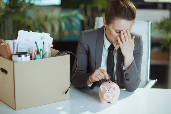 New job. sad modern female employee in modern green office in grey business suit with personal belongings in cardboard box putting coin into piggy bank.