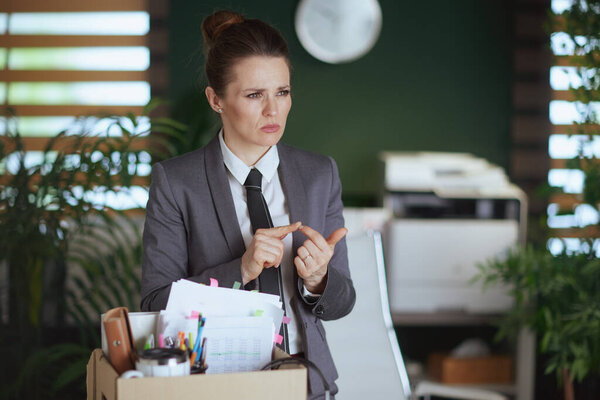 New job. pensive modern woman worker in modern green office in grey business suit with personal belongings in cardboard box.
