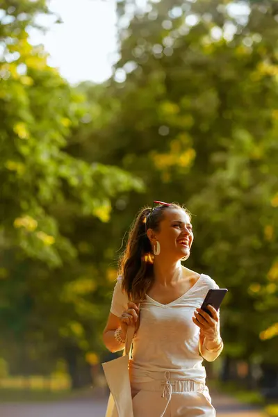 サマータイム スマートフォンを使ってトートバッグで白いシャツを着た幸せな若い女性と市立公園を歩く ロイヤリティフリーのストック画像