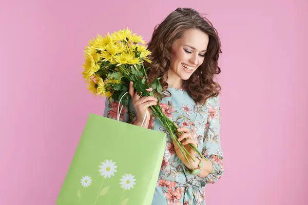 黄色の菊の花とピンクの背景に隔離された緑のショッピングバッグと花のドレスでスタイリッシュな女性を笑顔 ロイヤリティフリーのストック画像