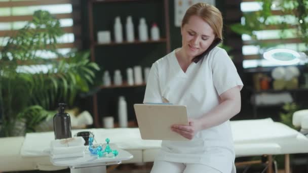 卫生保健时间 快乐的女性医疗按摩师在按摩室里用智能手机与剪贴板交谈 — 图库视频影像