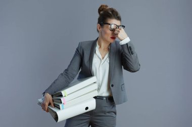 Gri arka planda gözlük ve klasörlerle gri takım elbiseli, orta yaşlı, moda bir kadın işçi..