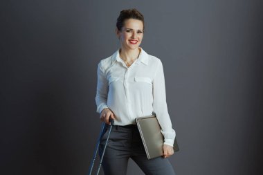 Gülümseyen, beyaz bluzlu, dizüstü bilgisayarlı ve tekerlekli çantalı modern iş kadını griye bürünmüş..