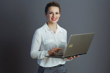 Beyaz bluzlu, gri arka planda dizüstü bilgisayarı olan zarif, küçük işletme sahibi bir kadın..