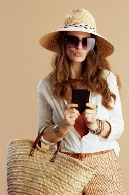 Plaj tatili Beyaz bluz içinde gülümseyen zarif bir kadın ve bej arka planda izole edilmiş şortlar, hasır çanta, hasır şapka ve cep telefonuyla selfie çeken güneş gözlükleri..