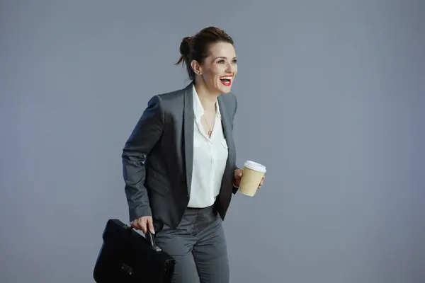 灰色の背景に対して走るコーヒー カップおよびブリーフケースとの灰色のスーツの現代中年の女性の従業員微笑 — ストック写真