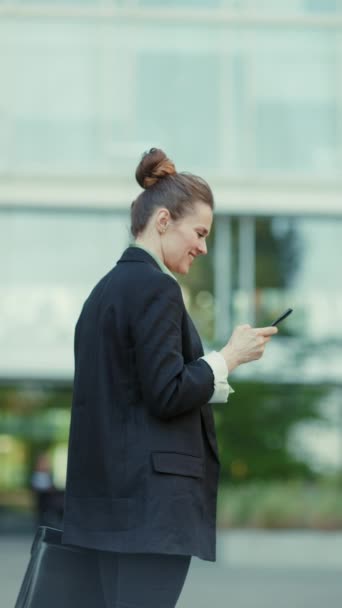 穿着黑色夹克 头戴智能手机 头戴手提箱走路的女商人站在写字楼附近 — 图库视频影像