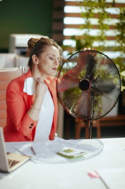 Sürdürülebilir iş yeri. İşyerindeki kırmızı ceketli, elektrikli yelpaze ve yaz sıcağından muzdarip peçeteli modern küçük işletme sahibi kadın..