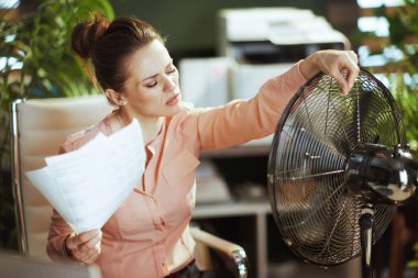 Sürdürülebilir iş yeri. Modern yeşil ofisteki 40 yaşında kadın çalışan. Belgeler ve elektrikli fan ile yaz sıcağından muzdarip..