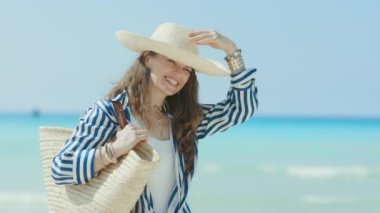 Gülümseyen şık orta yaşlı kadın okyanus kıyısında saman torbası ve hasır şapkayla.