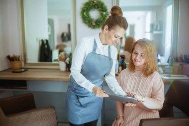 Modern saç stüdyosunda tableti ve müşterisi olan orta yaşlı kadın kuaför..
