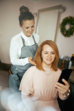 Modern güzellik salonundaki orta yaşlı kadın kuaför çalışanı ve akıllı telefonlu müşterisi..