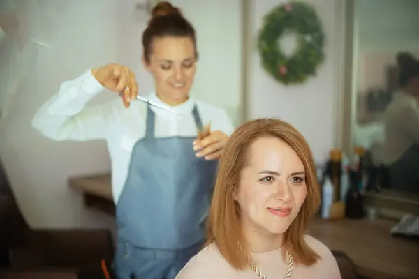 40岁的女性理发师在现代美容院用剪刀剪头发 免版税图库照片