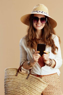 Plaj tatili gülümseyen orta yaşlı beyaz bluz ve şortlu ev kadını saman çantalı, akıllı telefonlu, yaz şapkalı ve güneş gözlüklü bej arka planda.