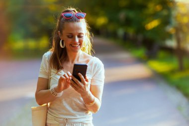 Yaz zamanı. Sırıtkan orta yaşlı beyaz tişörtlü, çantalı, akıllı telefon kullanan ve şehir parkında yürüyen bir kadın..