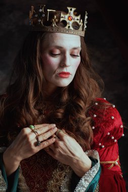 Kırmızı elbiseli, beyaz makyajlı ve tacı koyu gri arka planda olan ortaçağ kraliçesi..