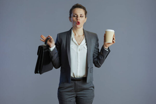stressed elegant 40 years old woman employee in grey suit with coffee cup and briefcase against gray background.