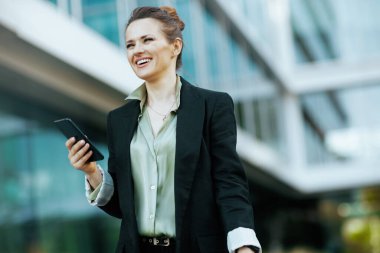 Gülümseyen modern 40 yaşındaki iş kadını ofis binası yakınında, akıllı telefon yürüyüşlü siyah ceketli..