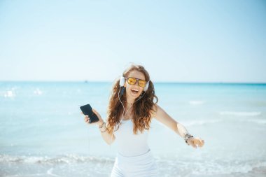 Okyanus kıyısındaki gülümseyen modern kadın akıllı telefonuyla dans ediyor ve kulaklıkla müzik dinliyor..