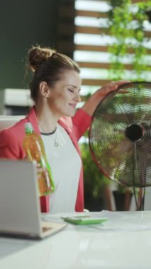 Sürdürülebilir iş yeri. Mutlu orta yaşlı muhasebeci kadın iş yerinde kırmızı ceketli, elinde bir şişe su ve elektrikli vantilatörle.