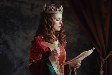 Üzerinde parşömen ve tacı olan koyu gri arka planda kırmızı elbiseli düşünceli ortaçağ kraliçesi..
