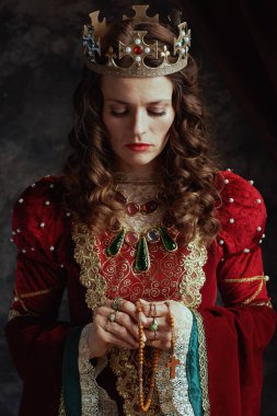 Ortaçağ kraliçesi kırmızı elbiseli, tespih ve tacıyla koyu gri arka planda dua ediyor..