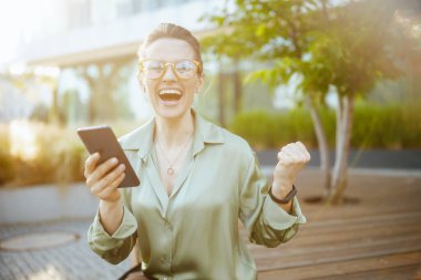 Mutlu orta yaşlı kadın çalışan ofis binasının yanında yeşil bluz ve gözlüklerle akıllı telefon ve neşe içinde bekliyor..