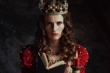 Kırmızı elbiseli, kitap, mum ve tacı koyu gri arka planda olan ortaçağ kraliçesi.