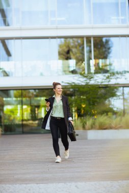 Orta yaşlı, gülümseyen, ofis binası yakınında, evrak çantalı, akıllı telefon kullanan ve yürüyen bir iş kadını portresi..