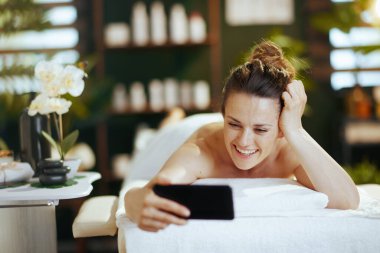 Sağlık hizmeti zamanı. Gülümseyen 40 yaşında bir kadın spa salonunda videodan konuşuyor ve masaj masasına uzanıyor..