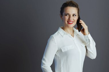 Beyaz bluzlu gülümseyen orta yaşlı iş kadını cep telefonuyla konuşuyor ve gri arka plana bakıyor..