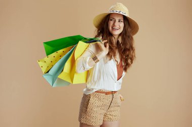 Plaj tatili Beyaz bluzlu ve şortlu mutlu orta yaşlı ev kadını bej arka planda izole edilmiş alışveriş çantası ve hasır şapkalı..