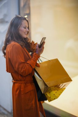 Merhaba Eylül. Mutlu modern 40 yaşındaki turuncu trençkotlu, alışveriş çantalı, akıllı telefonlu ve sonbahar sarısı yapraklı kadın şehrin yakınındaki dükkanda..