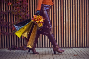 Merhaba Eylül. Kentte alışveriş torbaları ve sonbahar sarısı yaprakları olan kahverengi trençkotlu şık bir kadına yakın çekim..