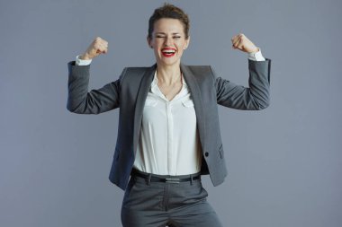 Gri takım elbiseli küçük işletme sahibi gülümseyen genç bir kadın gri üzerinde izole edilmiş pazılarını gösteriyor..