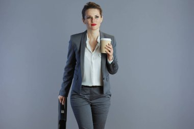 Gri takım elbiseli, kahve fincanı ve evrak çantasıyla gri arka planda yürüyen 40 yaşında küçük işletme sahibi bir kadın..